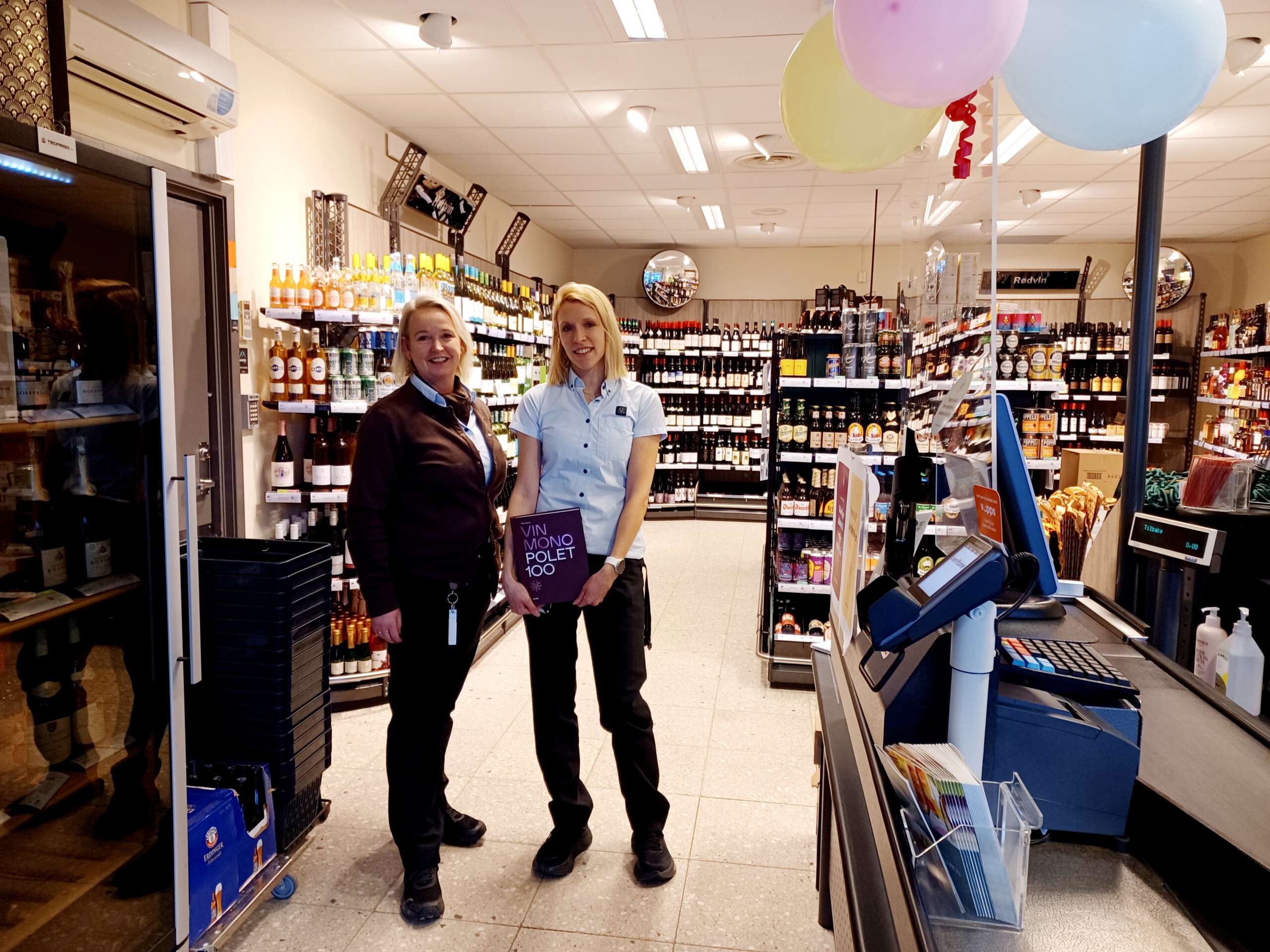 100 ÅR: Vinmonopolet i Norge har bursdag. I løten feires det med ballonger, kake og noe godt i glasset. I forbindelse med jubileet er boken "Vinmonopolet 100", av Olav Hamran gitt ut. Sjef Mona Kongsvoll Johansen og ekspeditør Tine Sellevold, har et eksemplar liggende i Løten. Her kan du lese om reisen gjennom det norske samfunnet, i hundre år. FOTO: Line Larsen